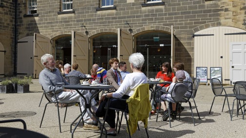 People are sat outside at tables and chairs, you can see drinks and some food on the table. Behind them is the Stables Cafe with its doors open